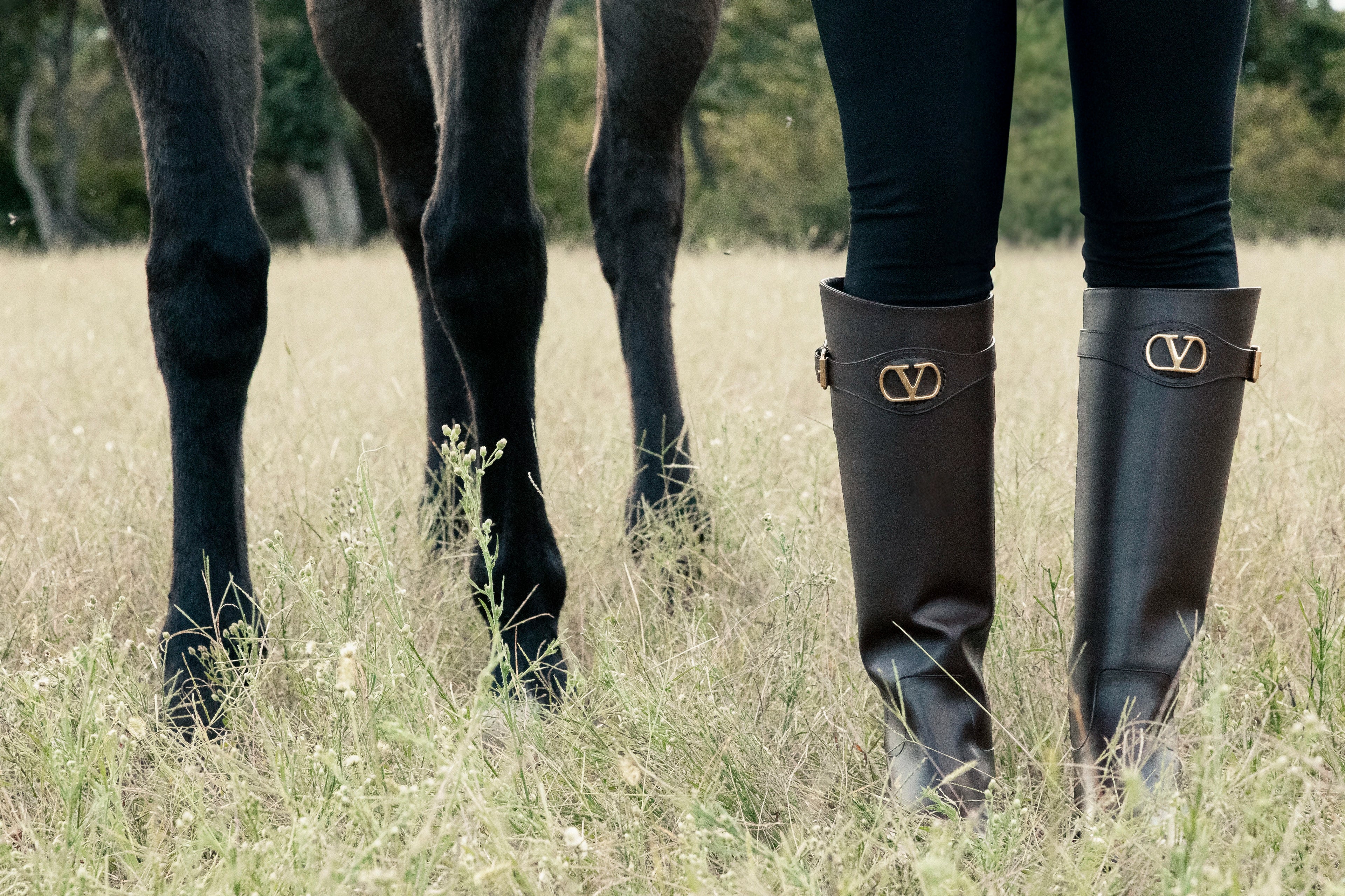 Bottes cavalières noires avec logo doré portées dans un champ aux côtés d’un cheval, illustrant la tendance équestre chic incontournable de la saison.
