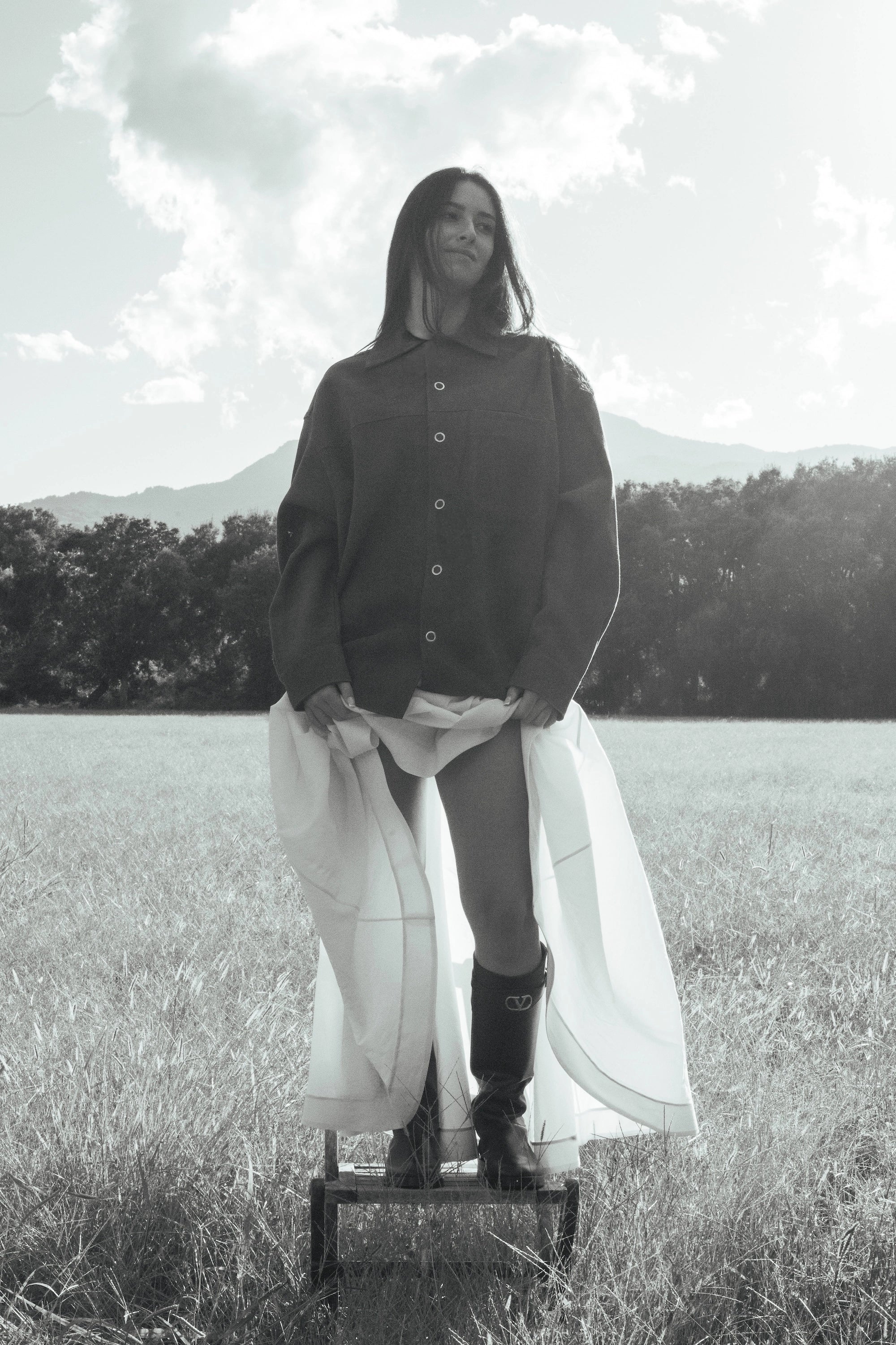 Woman standing in a field wearing an oversized dark jacket and Valentino boots, holding a white flowing coat, part of the myCompañero Women's Fall Winter 2025 collection.
