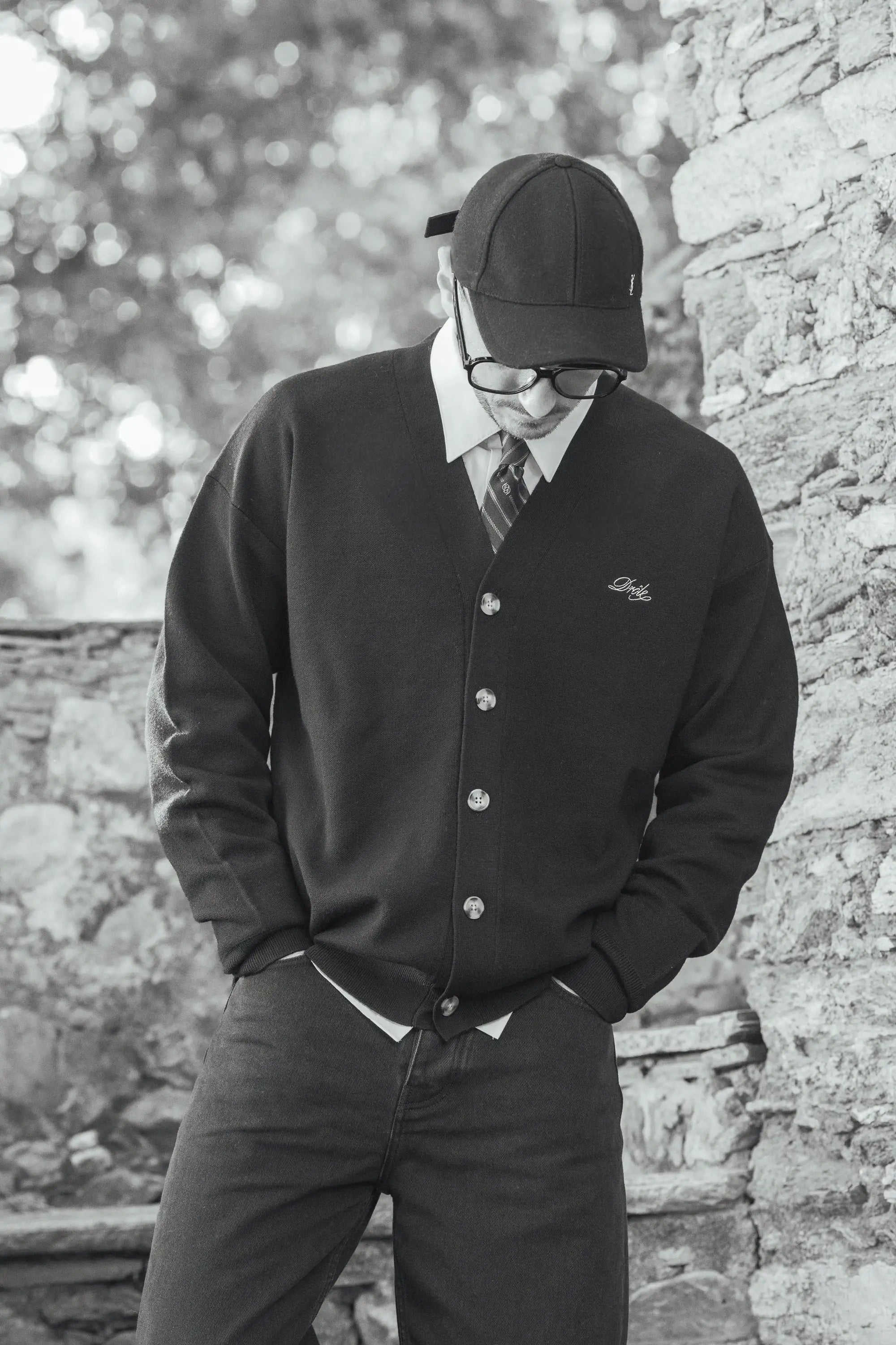Man wearing a black cardigan, white shirt, and tie with a cap and glasses, standing near a stone wall, part of the myCompañero Men's Fall Winter 2025 collection.