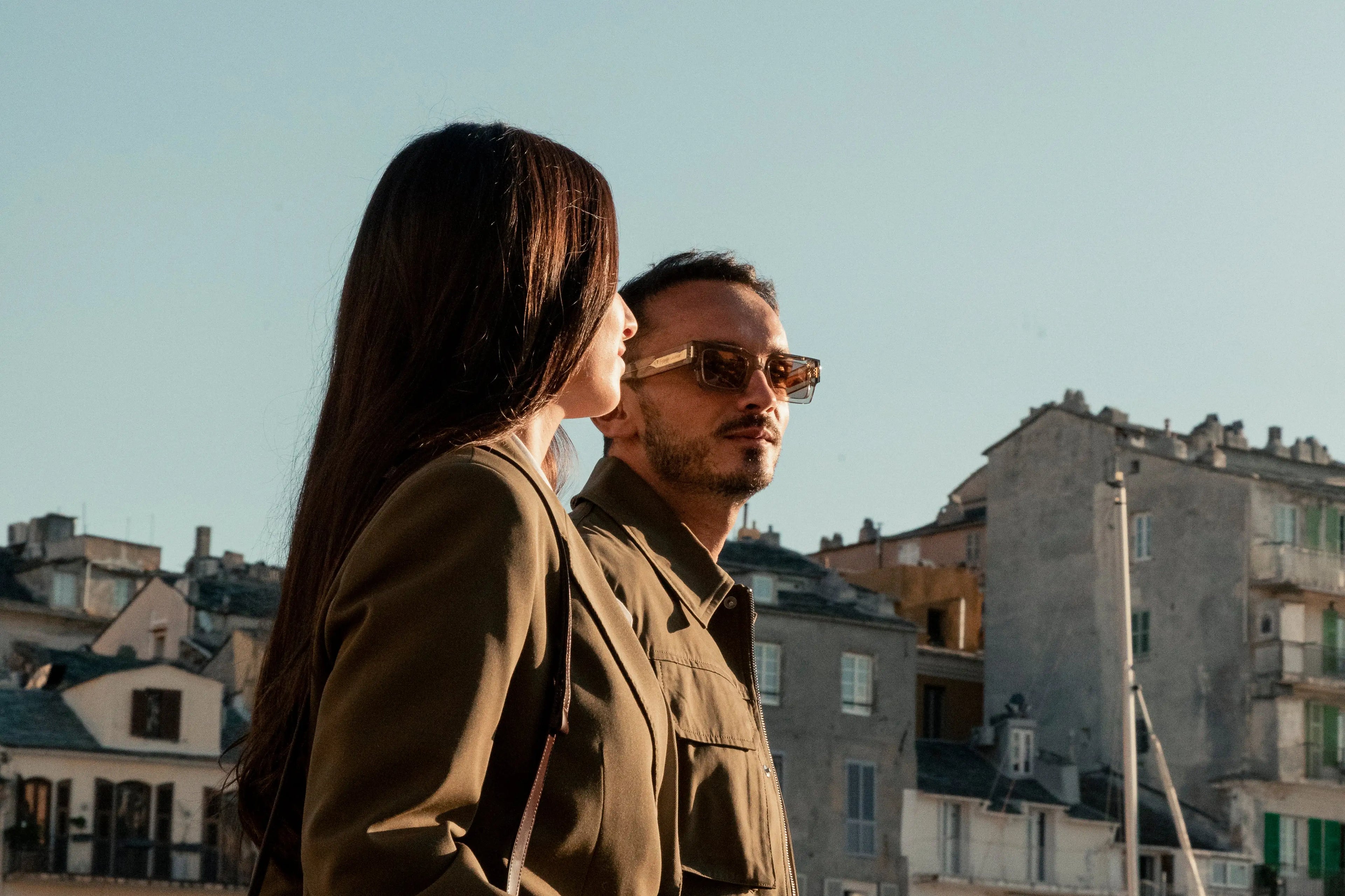Man and woman wearing khaki jackets and sunglasses in sunlight, standing in front of old Corsican buildings, representing the myCompañero New In Fall Winter 2025 campaign.